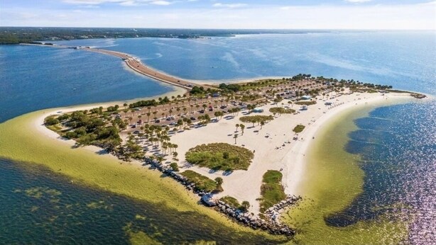 An aerial view of a coastal resort with a sandy beach, clear blue waters, and a small island with structures and greenery.