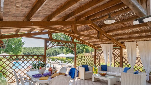 A covered outdoor seating area with wicker furniture, a table with food and drinks, and a view of a pool and greenery. - Belvilla by OYO Selvatello Laura Trilo Cinque (Casole d'Elsa)