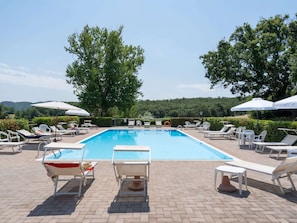 Ein rechteckiger Pool, umgeben von weißen Liegestühlen und Sonnenschirmen, mit Bäumen und einer hügeligen Landschaft im Hintergrund.