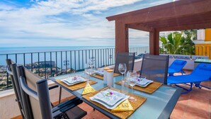 Mesa de jantar posta para dois, com vista desobstruída para o mar e uma cidade costeira ao fundo.