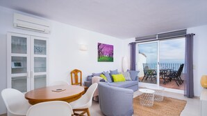 Uma sala de estar moderna com um sofá cinzento, uma mesa de jantar de madeira com cadeiras brancas, um aparador de vidro e uma varanda com vista.