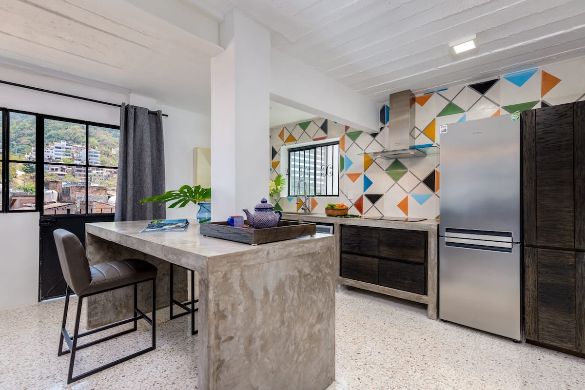 Une cuisine moderne dotée d’un îlot central, de tabourets de bar, d’un réfrigérateur en acier inoxydable et d’un revêtement mural géométrique aux couleurs vives.