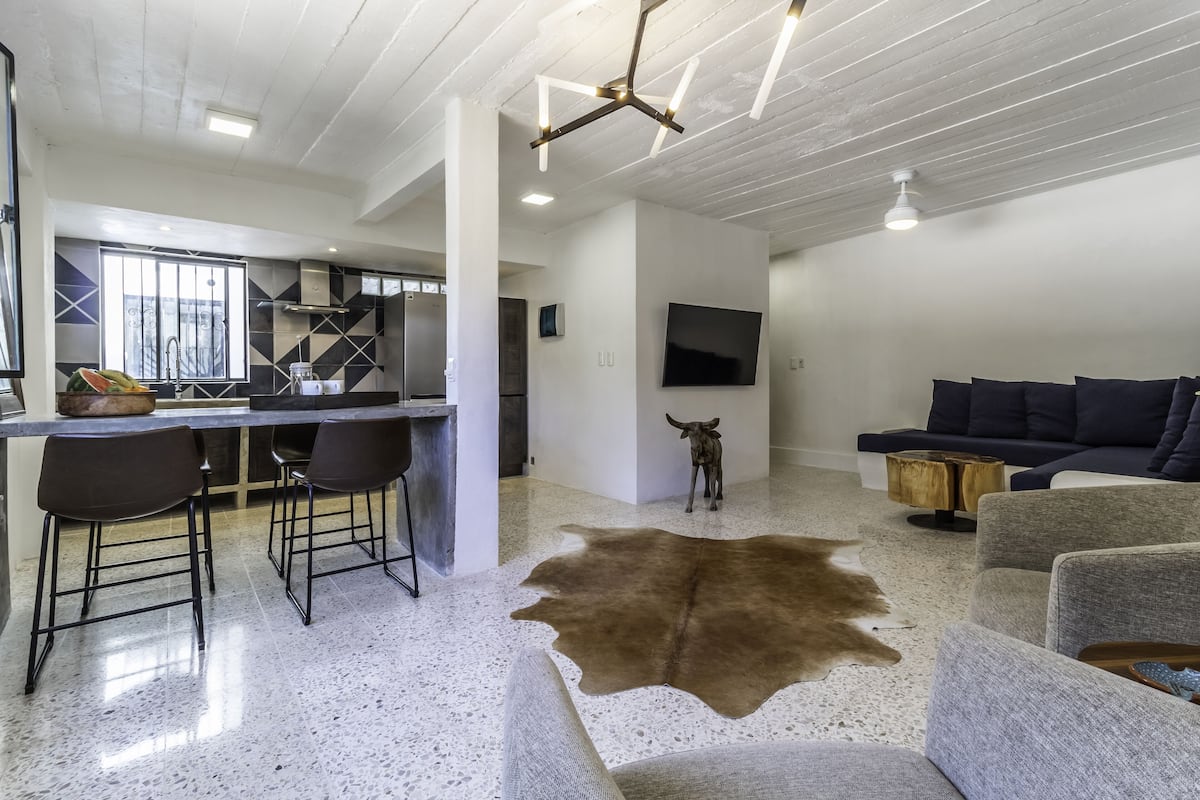 Un salon moderne avec un grand tapis en cuir, un coin repas doté de tabourets de bar, une cuisine avec un réfrigérateur en acier inoxydable et un téléviseur fixé au mur.