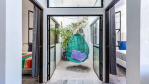 Een smalle gang met een hangstoel, een bed met kleurrijk beddengoed en een potplant.