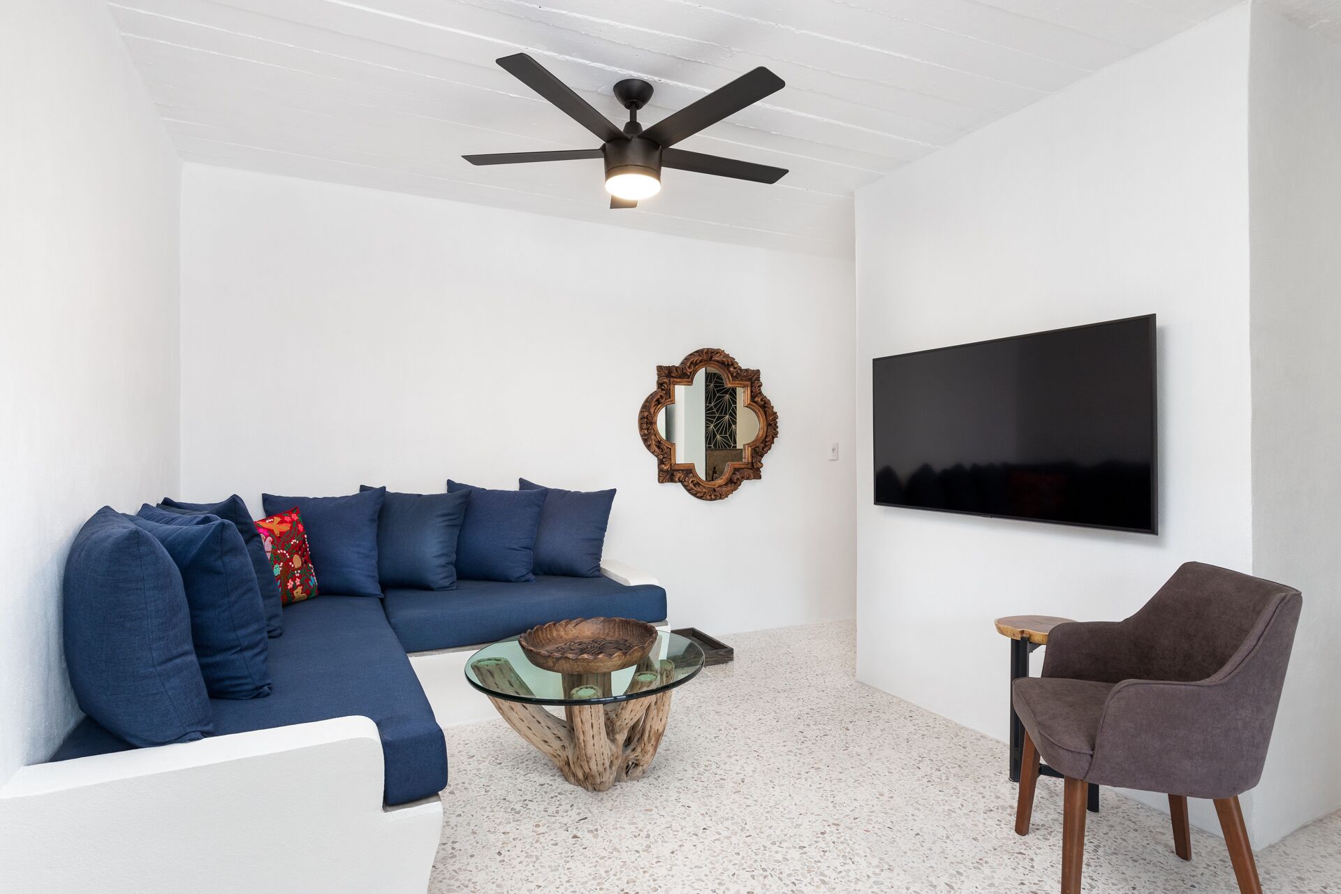 Ein modernes Wohnzimmer mit einem blauen Sofa, einem Glastisch, einem Holznachttisch, einem braunen Sessel, einem Deckenventilator, einem wandmontierten Fernseher und einem dekorativen Spiegel.