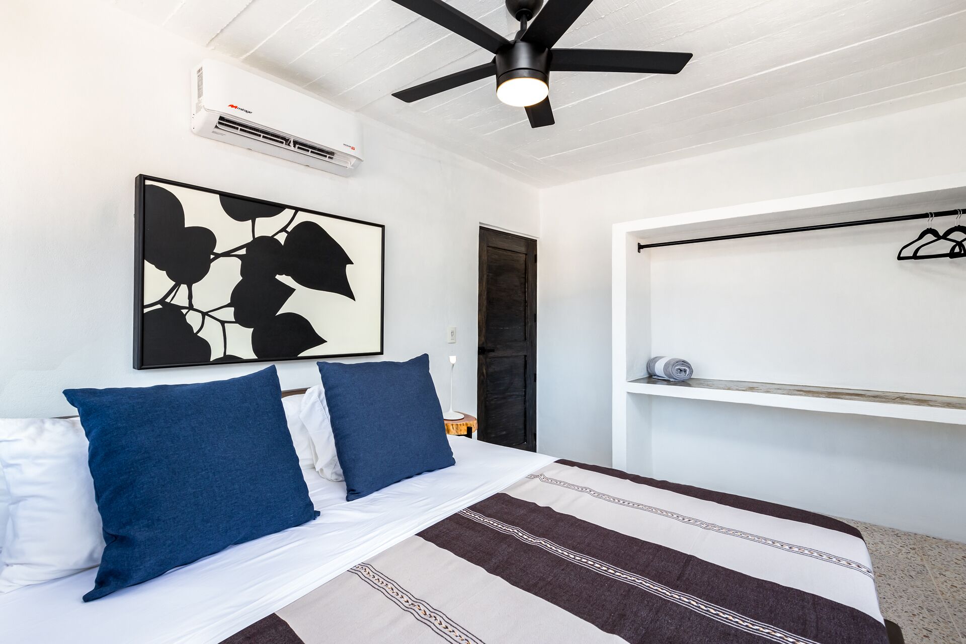 Une chambre à coucher comprenant un lit, un ventilateur de plafond, un climatiseur mural, une œuvre d’art abstraite encadrée et un portant à vêtements.