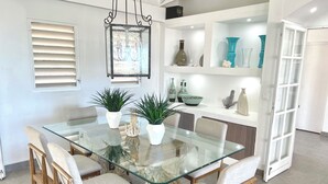 Un coin repas avec une table en verre entourée de chaises en bois, de plantes en pot et d’objets décoratifs disposés sur des étagères.