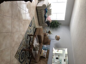 Un salon avec un tapis à motifs, un canapé marron, un fauteuil blanc et une table basse. On y trouve une fenêtre avec des stores, une plante en pot et une lampe sur un pied.
