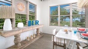A cozy outdoor dining area with a wooden bench, a table set for two, and a view of the outdoors through large windows with blue shutters. - LAGON BLANC (LE VAUCLIN)