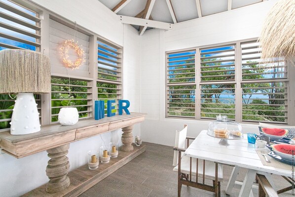 A cozy outdoor dining area with a wooden bench, a table set for two, and a view of the outdoors through large windows with blue shutters.