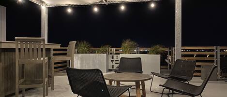 An outdoor patio area with a round table and four chairs, a wooden bench, and a concrete planter with plants.