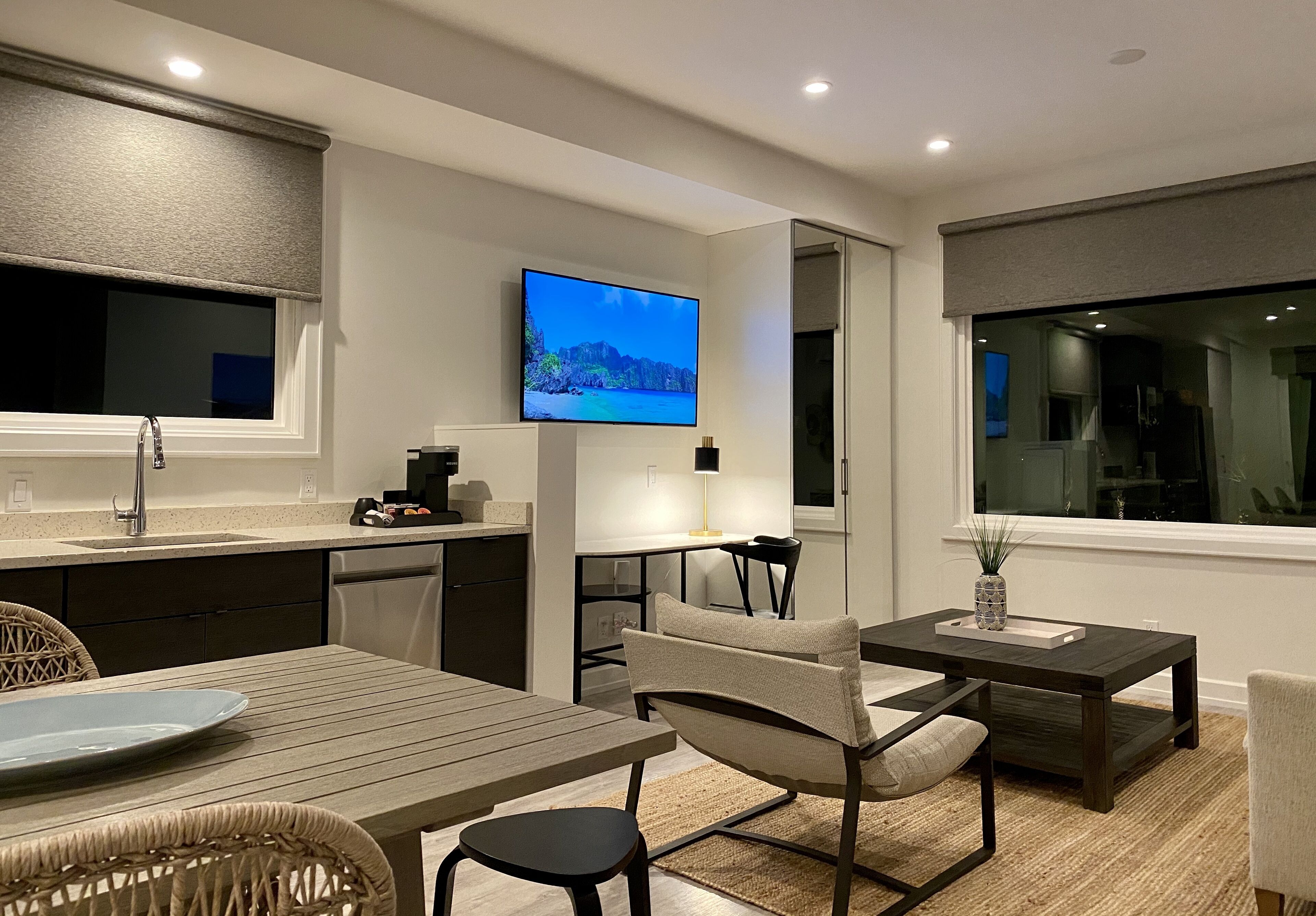 A modern kitchen with a flat-screen TV, a dining area with a table and chairs, and a view of the outdoors through a window.