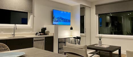A modern kitchen with a flat-screen TV, a dining area with a table and chairs, and a view of the outdoors through a window.