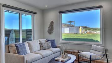 A modern living room with a sofa, a coffee table, and a chair. There are large windows offering a view of the outdoors.