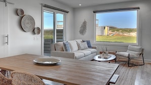 A modern living room with a wooden dining table, a sofa, and a chair. There are large windows with a view of the outdoors. - Lively Beach (Corpus Christi)
