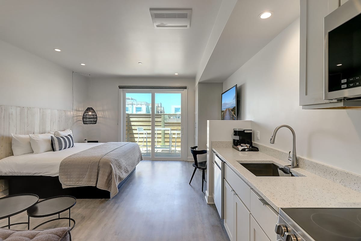 Una habitación de hotel moderna con cama, una pequeña mesa de comedor, una kitchenette y un amplio ventanal con vista a la ciudad.