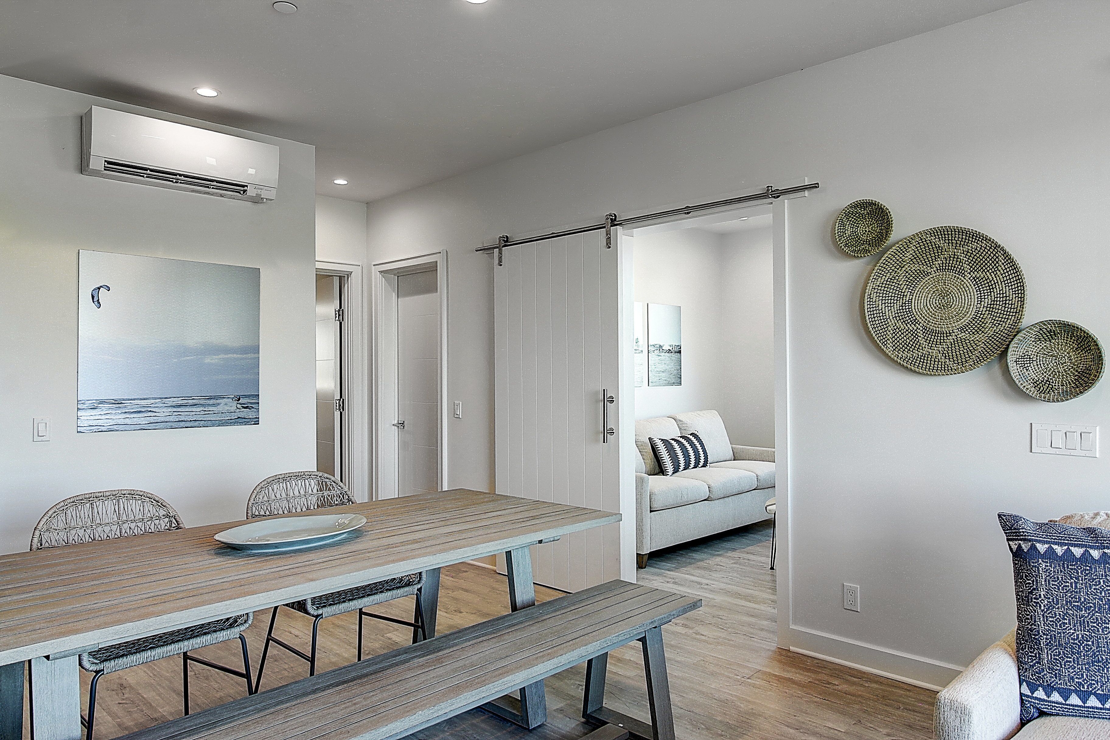 A modern dining area with a wooden table and bench, a view of the ocean through a window, and a hallway leading to a living area with a sofa and decorative plates on the wall.