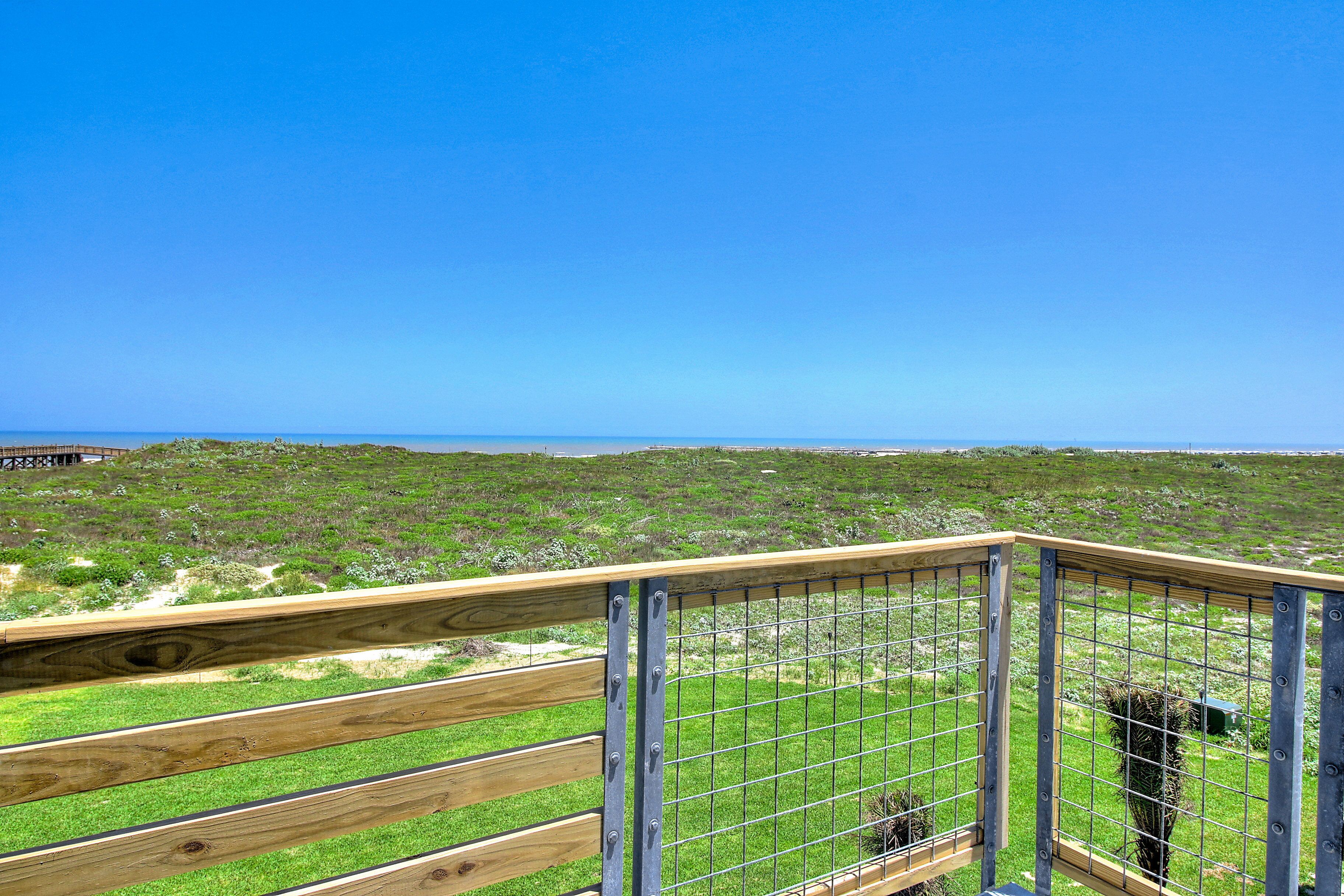 Une terrasse en bois avec une rambarde en métal, donnant sur un paysage verdoyant sous un ciel bleu dégagé.