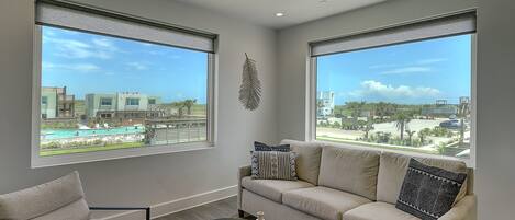 A modern living room with a beige sofa, a matching armchair, and a coffee table with a tray. Large windows offer a view of a pool and buildings.