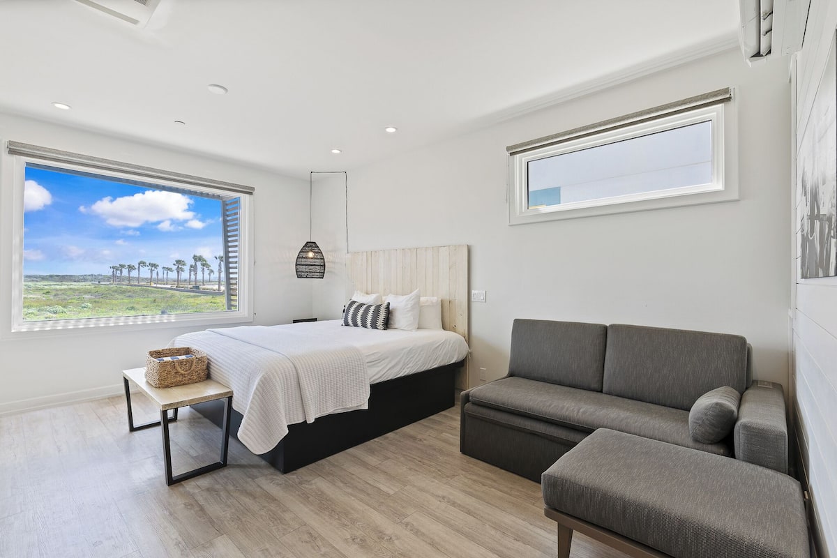 Un dormitorio moderno con cama, sofá gris, una mesita pequeña y un ventanal con vistas a palmeras.