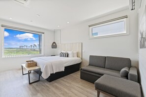 Une chambre moderne avec un lit, un canapé gris, une petite table et une grande fenêtre donnant sur des palmiers.