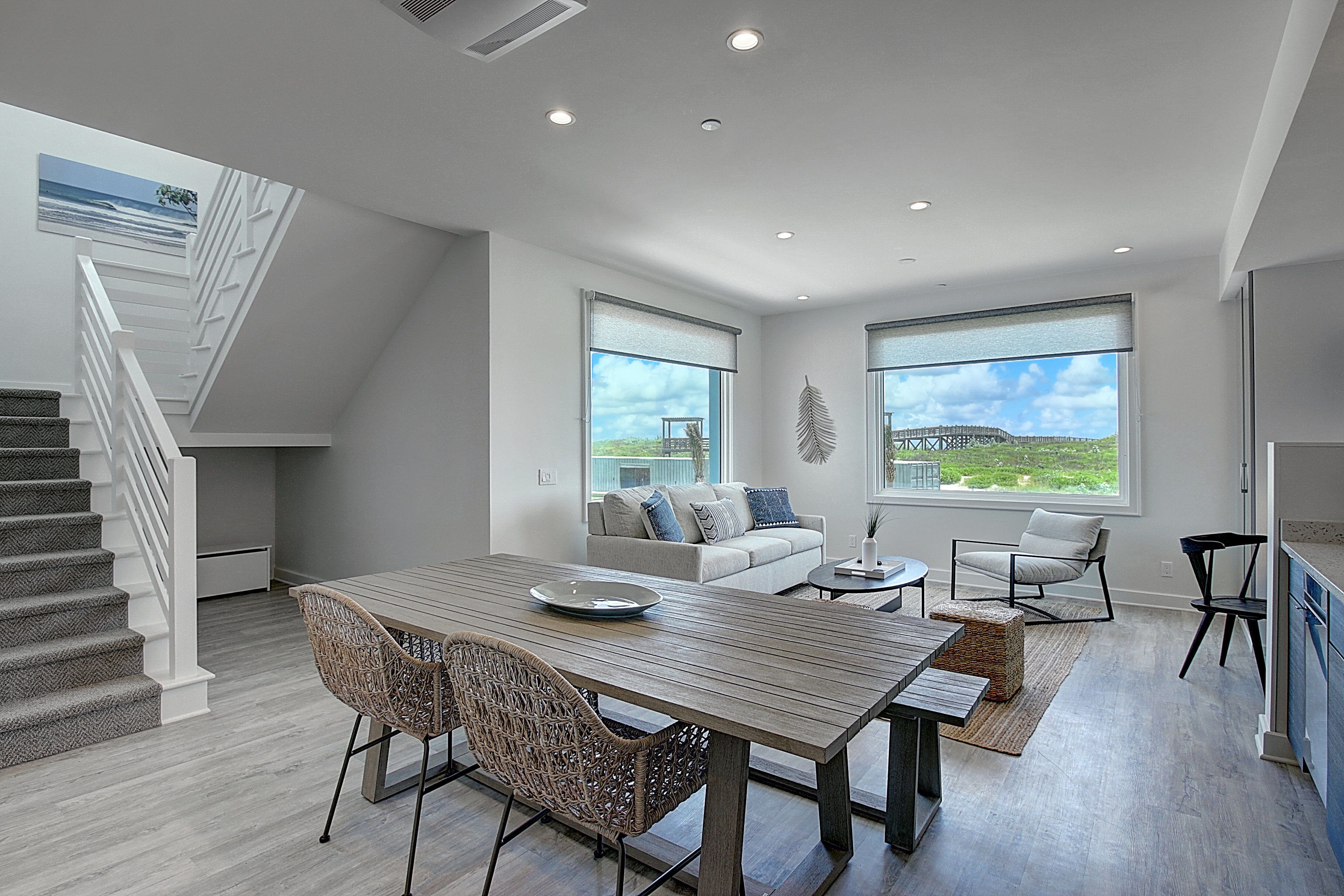 A modern living room with a dining area, a staircase, and large windows offering a view of the outdoors.