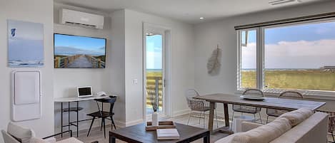 A modern living room with a large window, a flat-screen TV, a laptop, and a dining area with a wooden table and chairs.