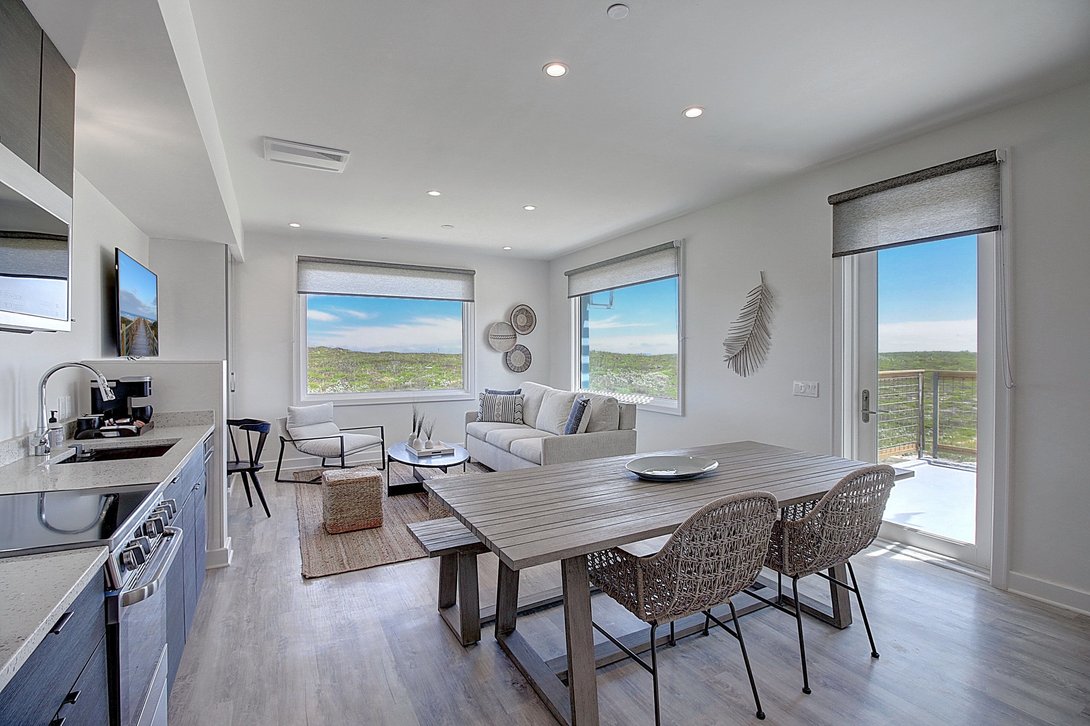 A modern kitchen with a built-in oven and sink, a dining area with a wooden table and wicker chairs, a living room with a sofa and a large window overlooking a landscape.
