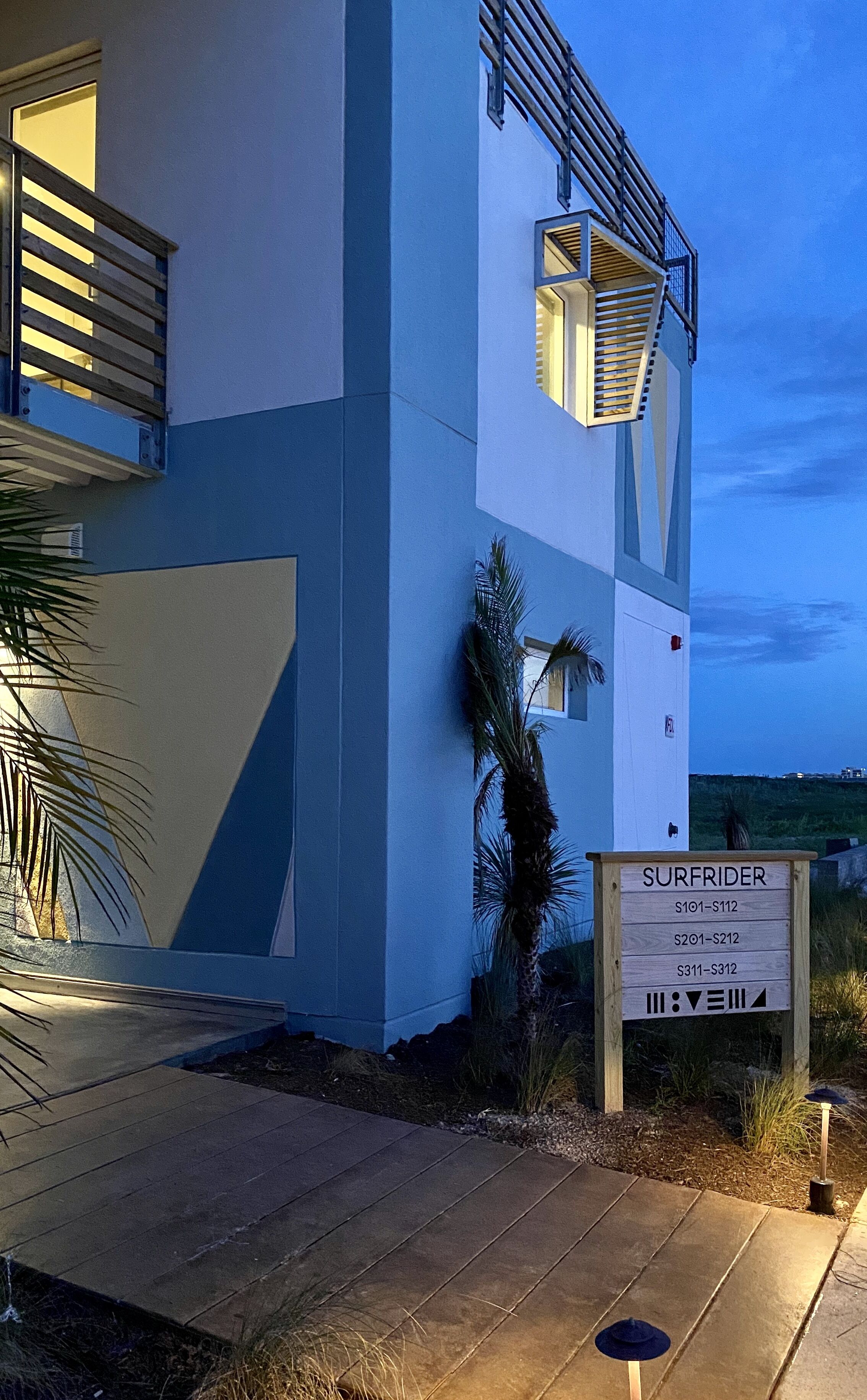 Un bâtiment moderne à la façade bleue et blanche, une enseigne indiquant « SURFRIDER » avec les numéros des chambres, et un palmier au premier plan.
