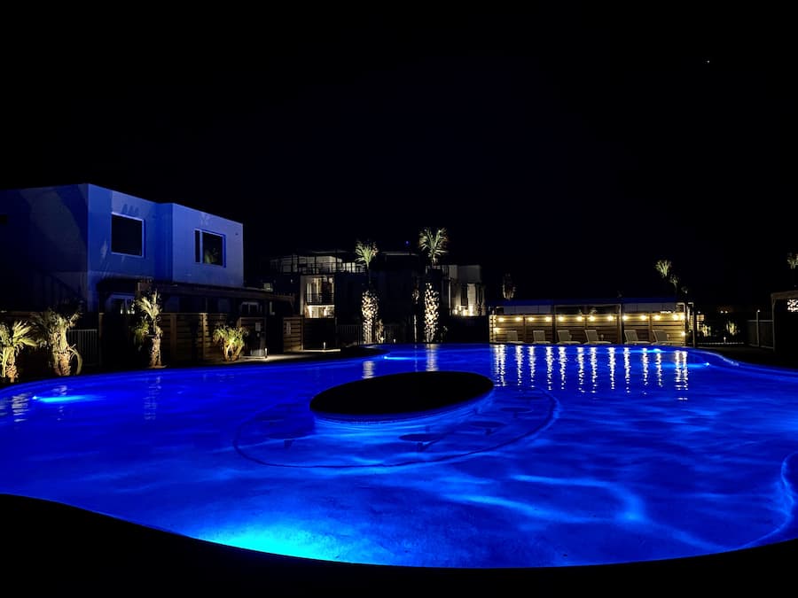 Una piscina con borde iluminado en azul, rodeada de edificios modernos y palmeras.