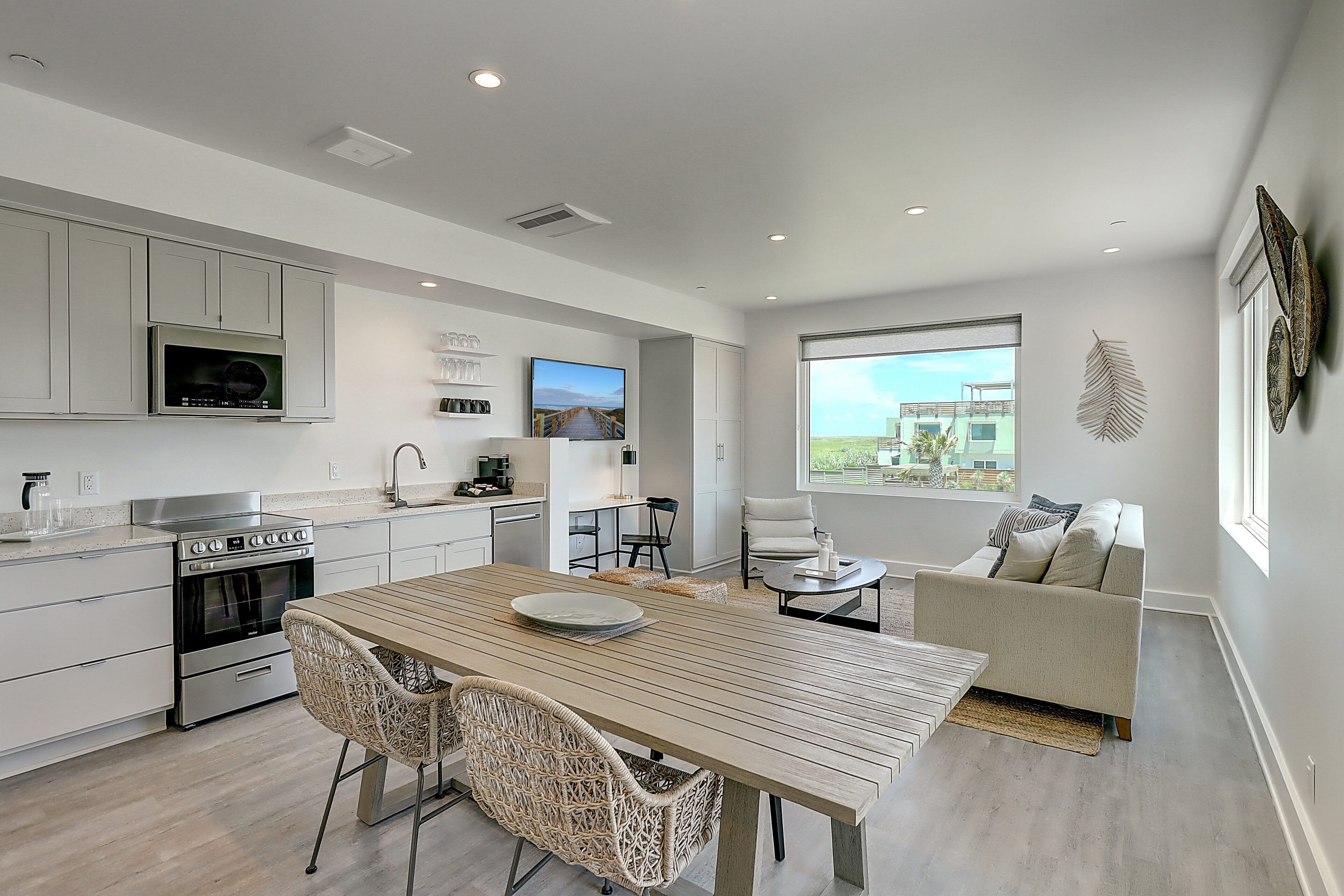 A modern kitchen with built-in appliances, a dining area with a wooden table and wicker chairs, and a living area with a sofa and a large window offering a view.