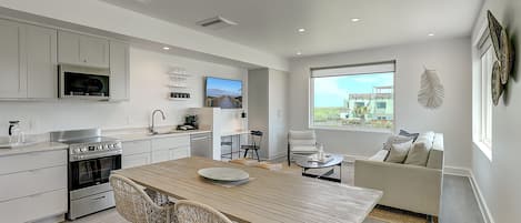 A modern kitchen with built-in appliances, a dining area with a wooden table and wicker chairs, and a living area with a sofa and a large window offering a view.