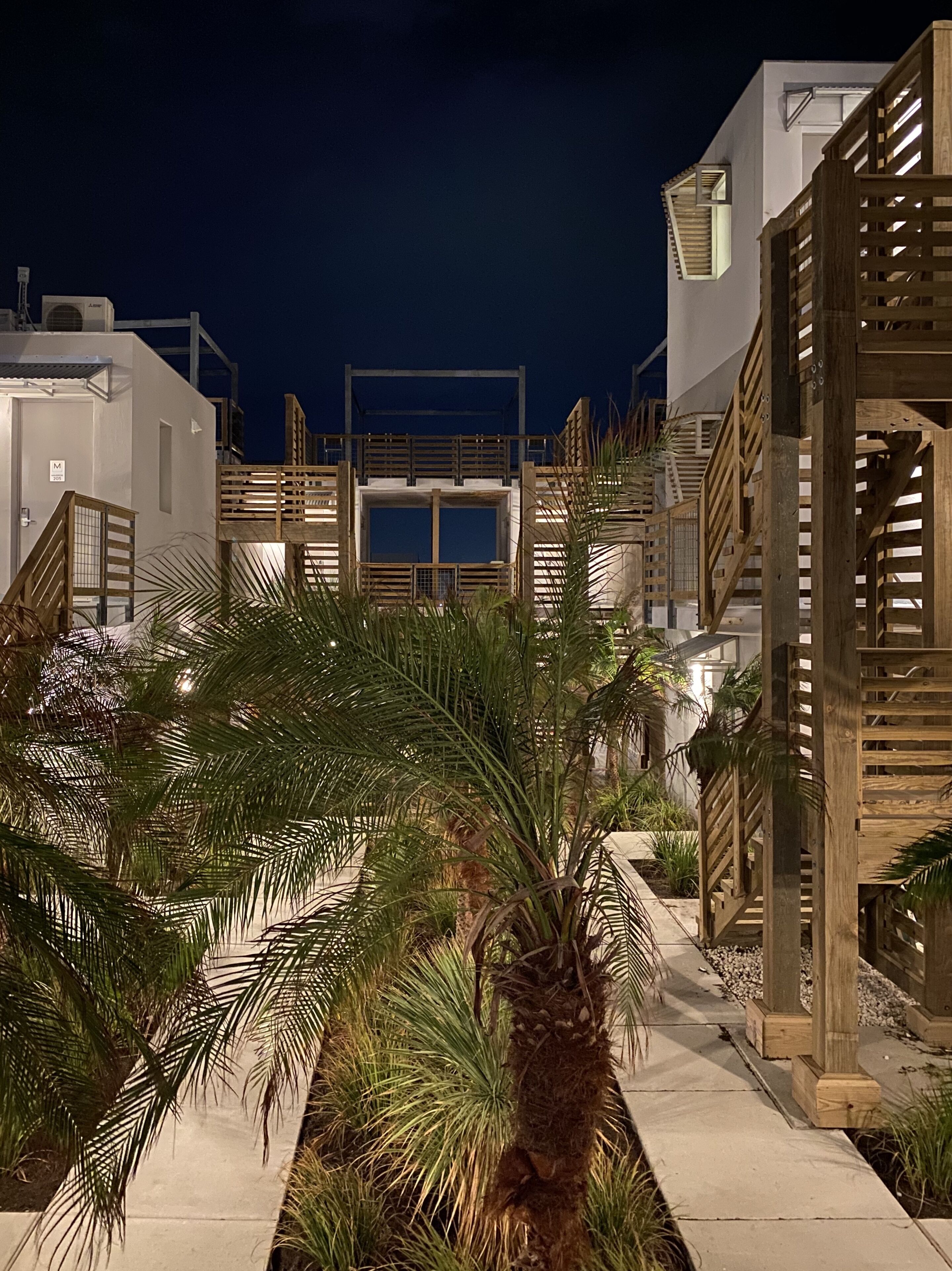 Vista nocturna de un edificio moderno con terraza de madera y palmeras.