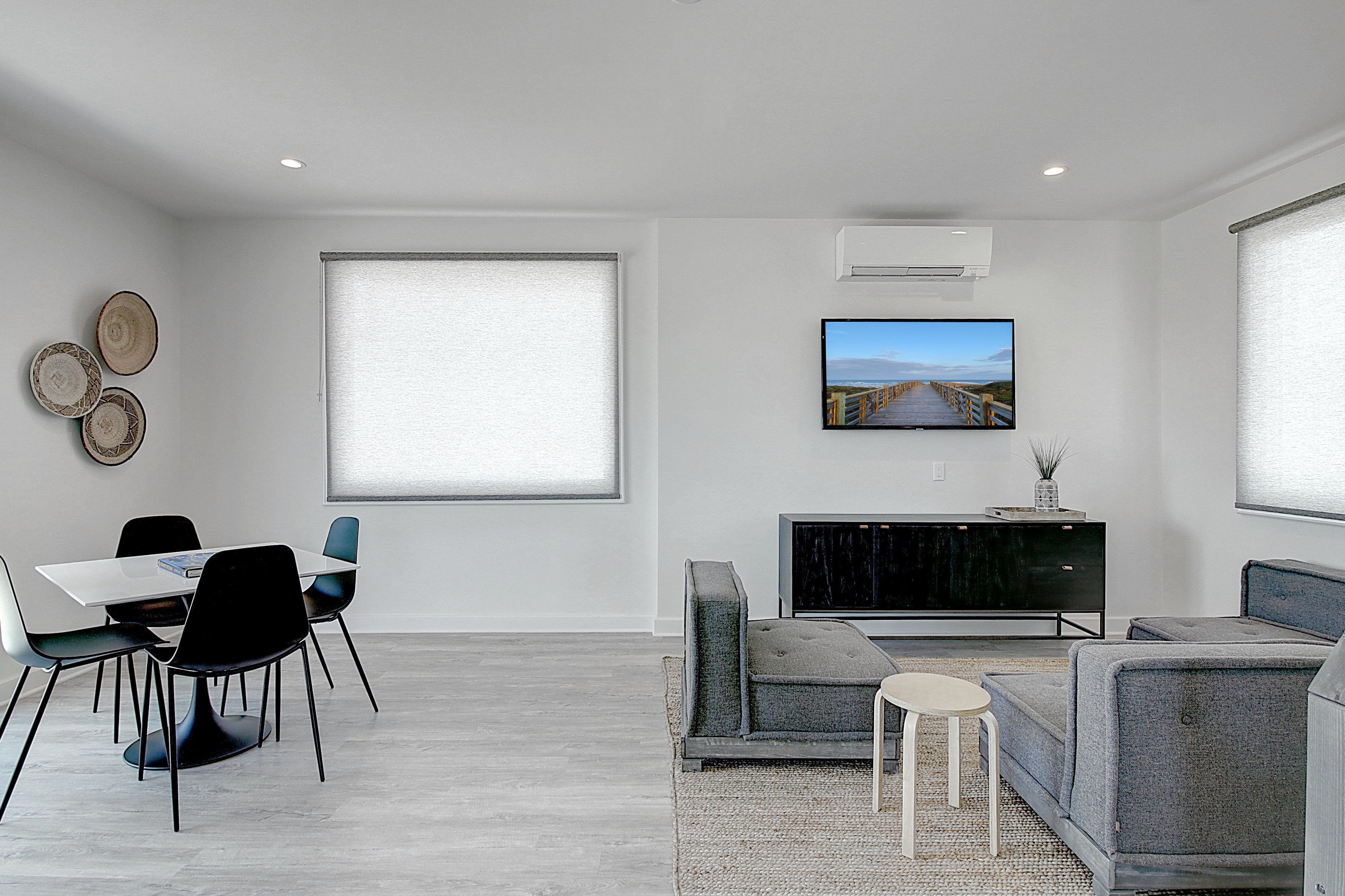 A modern living room with a dining area, a flat-screen TV mounted on the wall, and a large window with blinds.