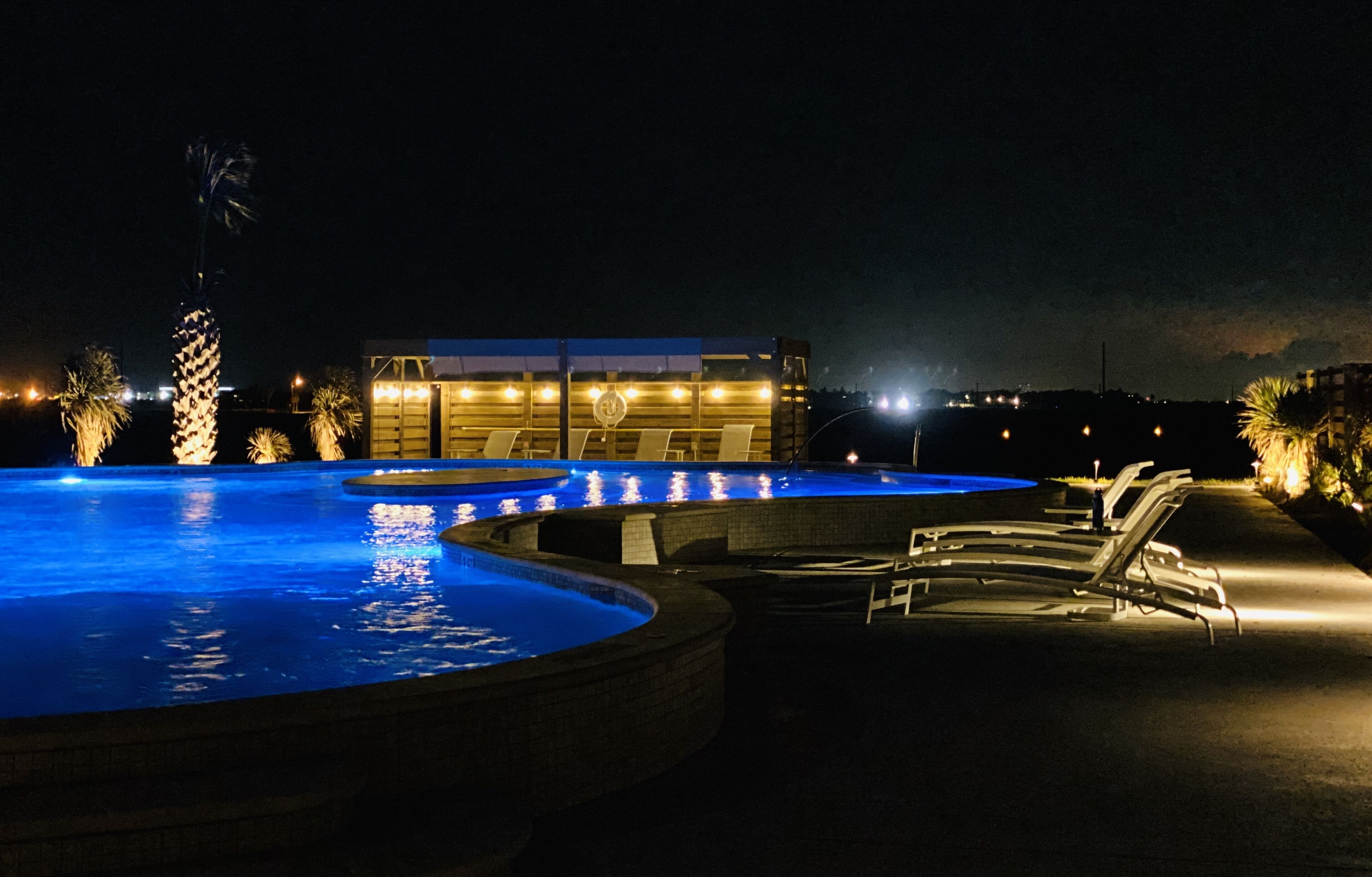 Zona de piscina iluminada por la noche con sillas de descanso y un edificio al fondo.