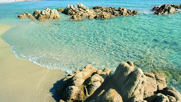 Ein Strand mit klarem türkisfarbenem Wasser, Felsformationen und einem sandigen Ufer.