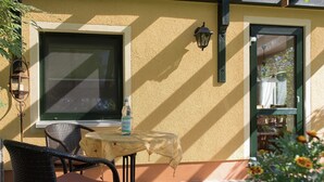 A patio with a wicker chair, a small table, a bottle of water, and a window with a view of the garden. - Apartment in Saxony With Terrace (Crottendorf)