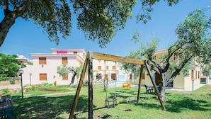 A playground with swings and a small table, surrounded by grass and trees, with a building in the background.