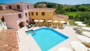 A swimming pool with sun loungers and umbrellas, surrounded by buildings with terracotta roofs.