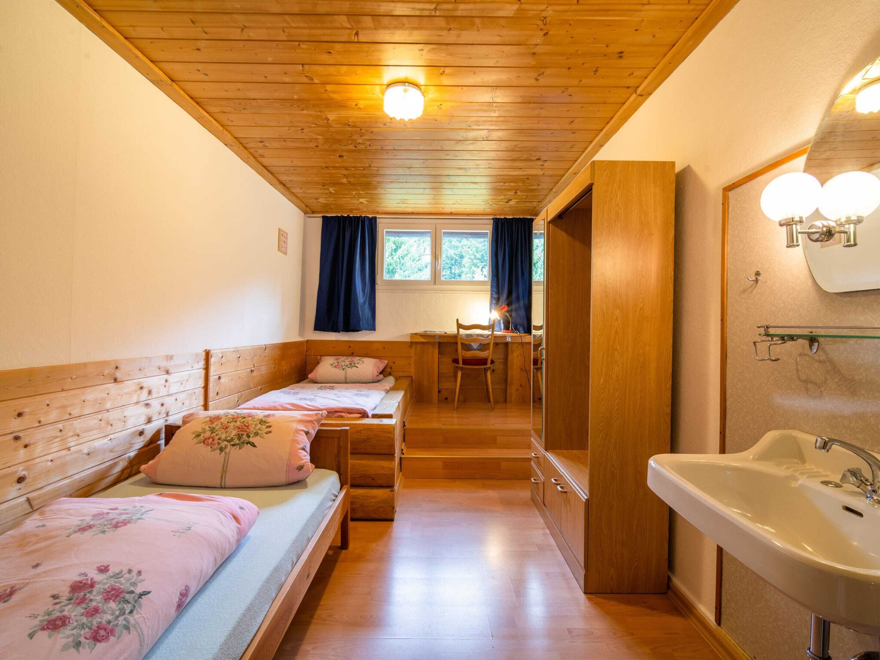 A cozy room with wooden floors, a bed with floral bedding, a desk with a chair, and a bathroom with a sink and mirror.