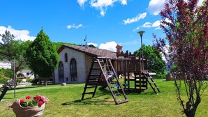 A playground with a wooden structure, a large potted plant, and a building with arched windows in the background. - Family Cottage in Cagli With Pool (Cagli)