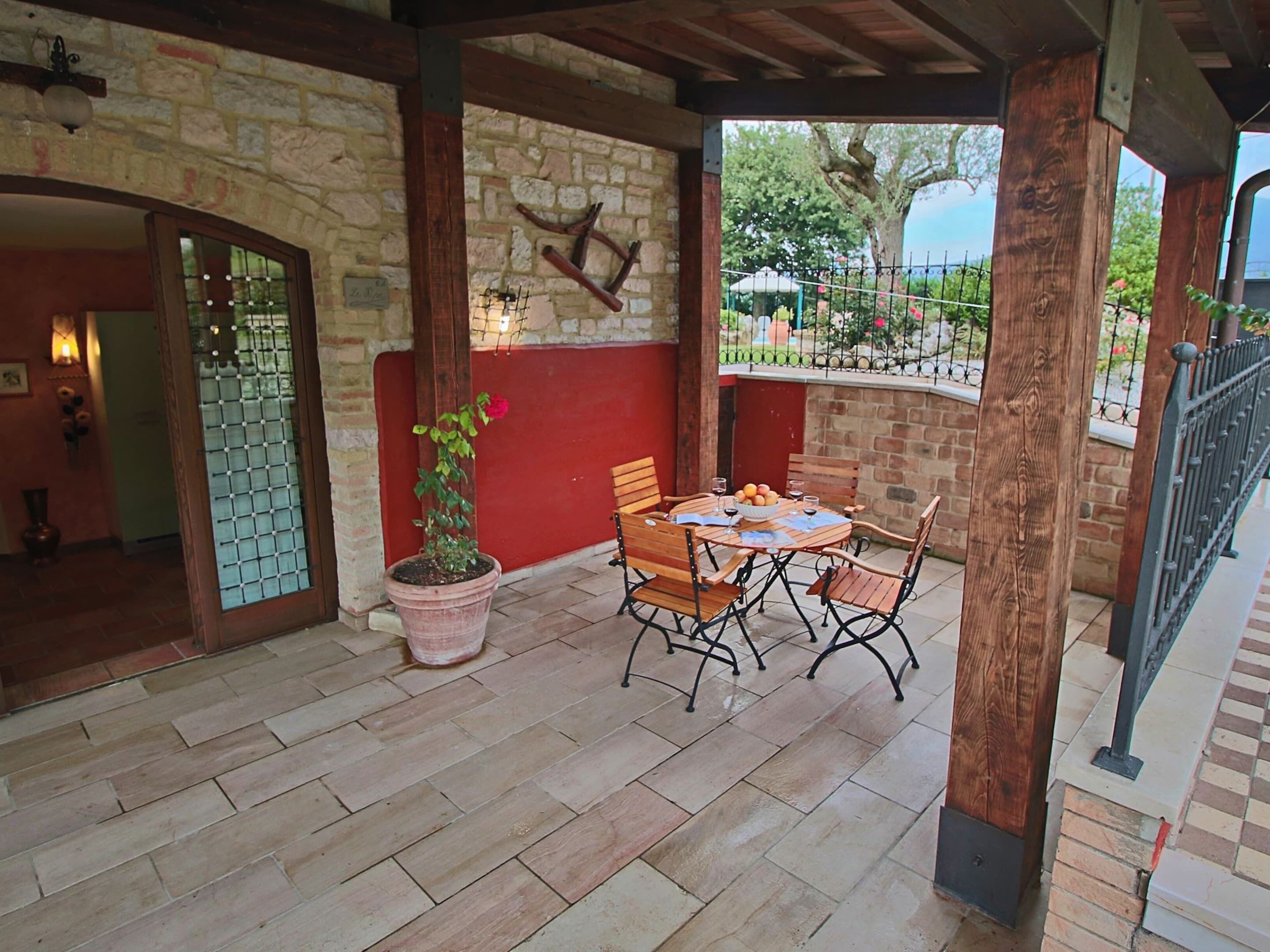 A covered patio with a stone wall, wooden beams, a table with chairs, a potted plant, and a view of the garden.