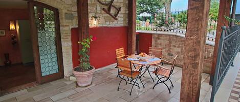 A covered patio with a stone wall, wooden beams, a table with chairs, a potted plant, and a view of the garden.