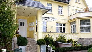 Un edificio de dos pisos con un porche cubierto, una escalera que conduce a la entrada y una pequeña zona exterior con mesa y sillas.