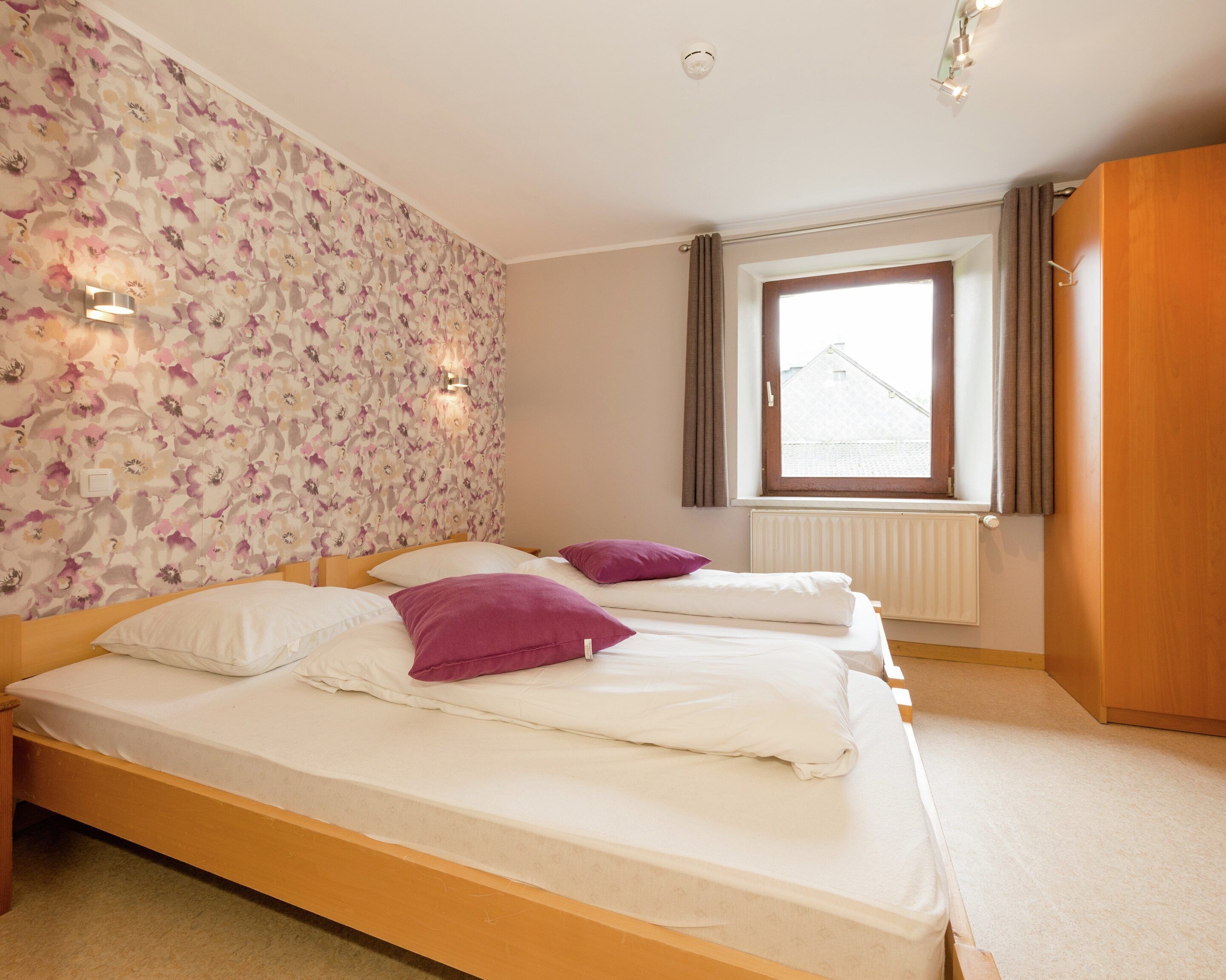 A bedroom with two beds, a window with curtains, a radiator, and a floral wallpaper.