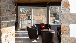 A room with stone walls, a black leather sofa, a round table, and a view of a fenced outdoor area. - Holiday Home with Sauna, Garden, Terrace, BBQ & Fireplace (Waimes)