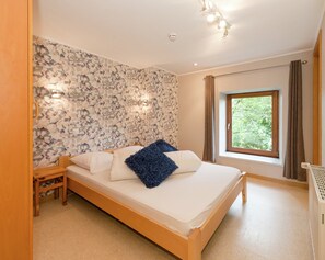 A bedroom with a bed, wooden headboard, a nightstand, a window with a view of greenery, and a wall with a floral pattern. - Holiday Home with Sauna, Garden, Terrace, BBQ & Fireplace (Waimes)