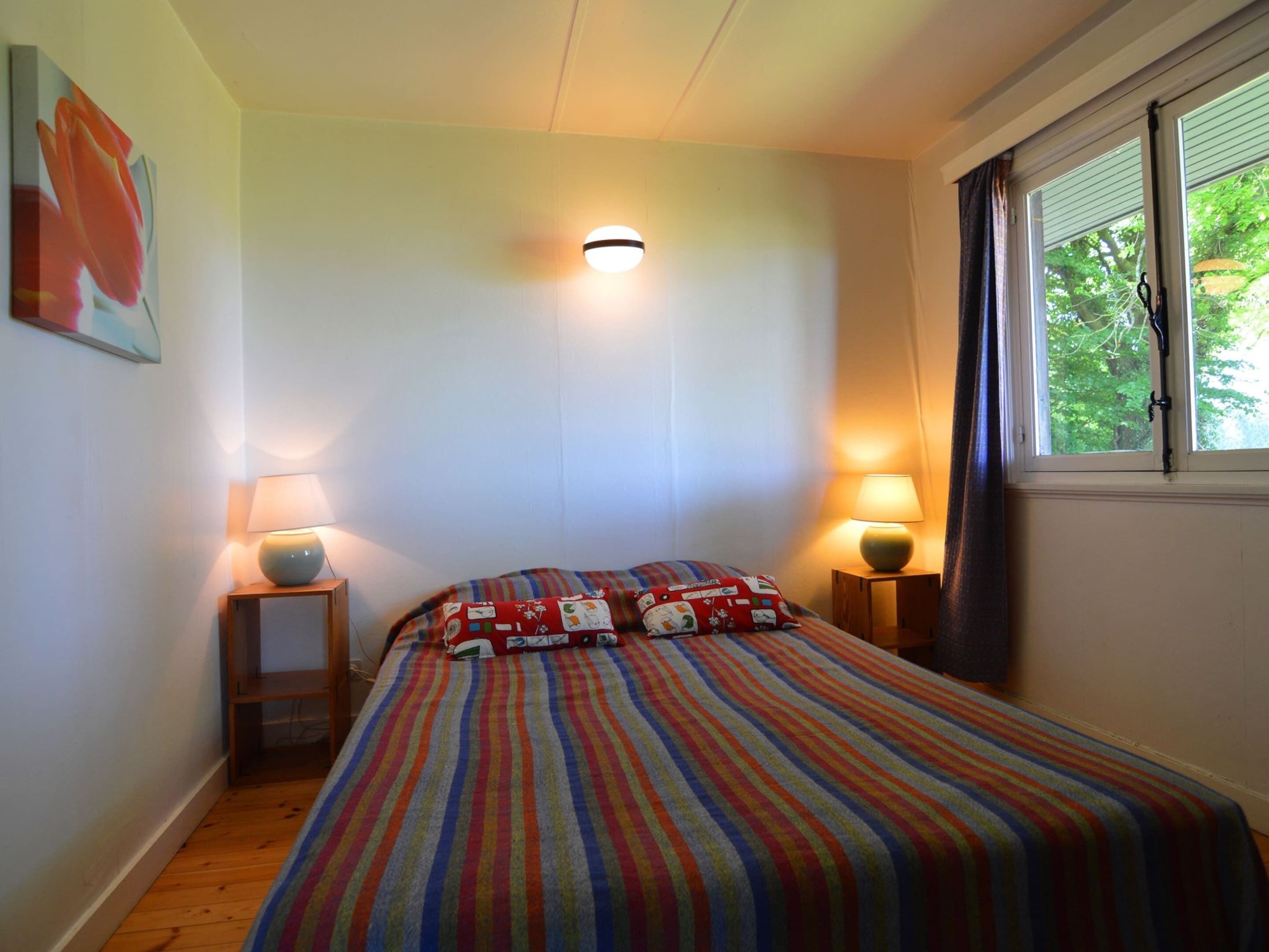 A bedroom with a striped bedspread, two bedside lamps, a wall-mounted light, and a window with curtains.