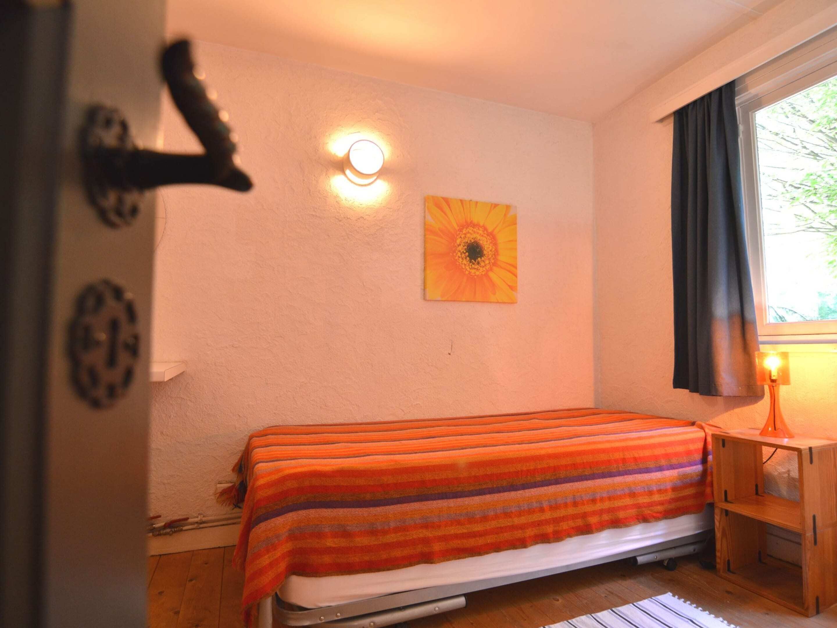 A small, cozy room with a single bed, a wall-mounted light, a window with curtains, and a wooden nightstand.
