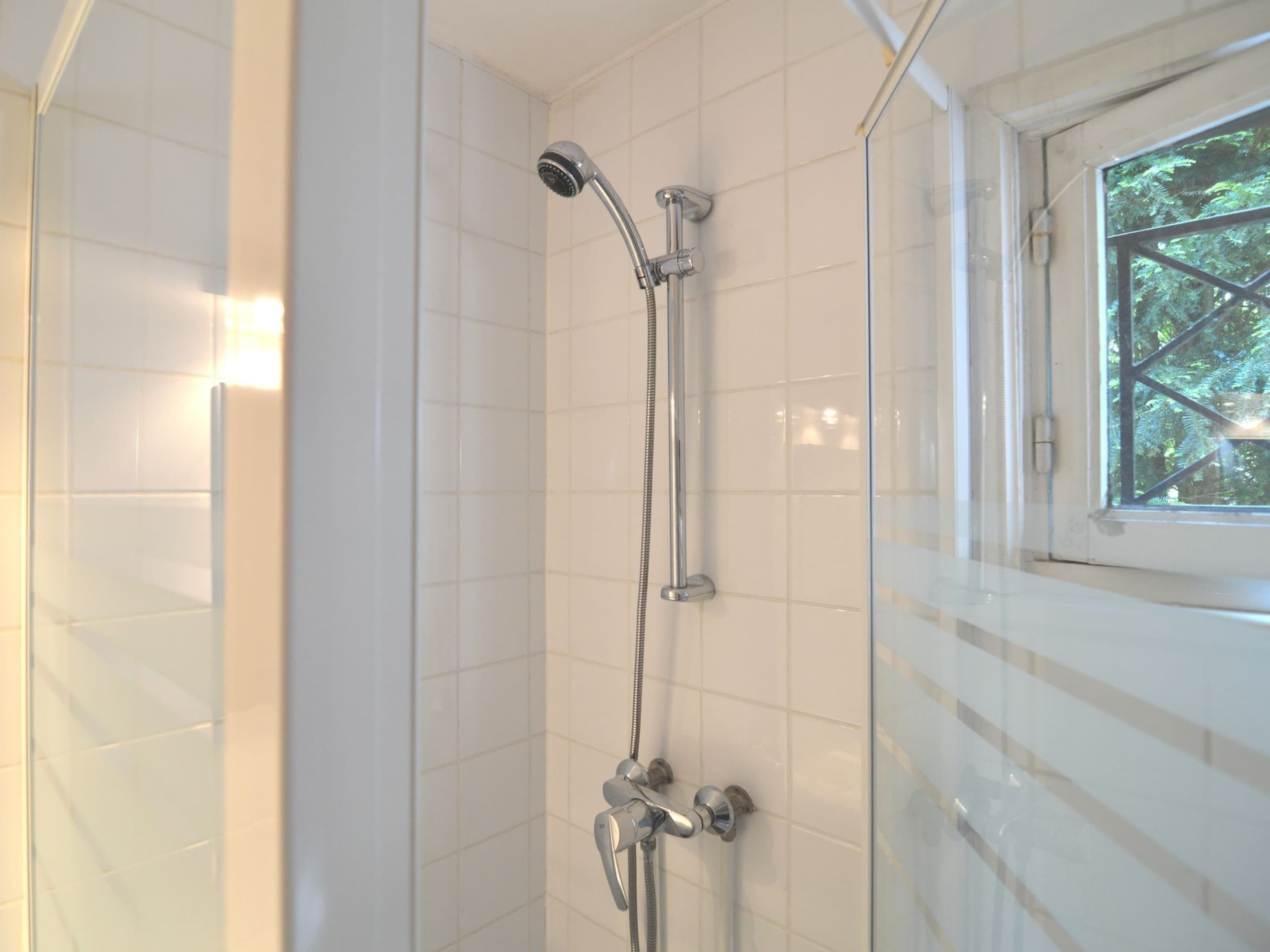 A shower with a handheld showerhead and a wall-mounted faucet, tiled walls, and a window with a view of trees.